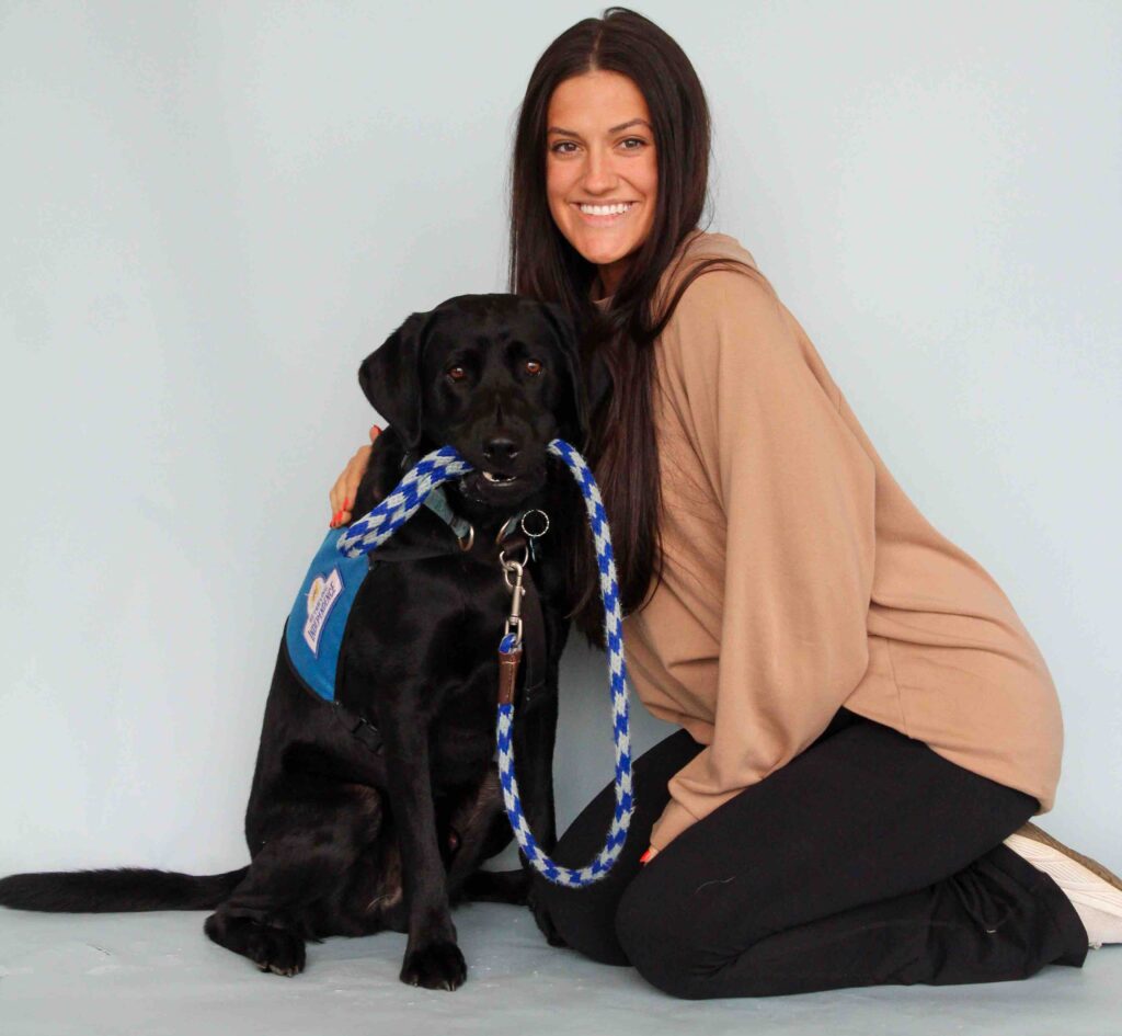 A happy volunteer sits next to a black labrador service dog who is holding their own leash in their mouth. Read to learn more about how to volunteer with RI.
