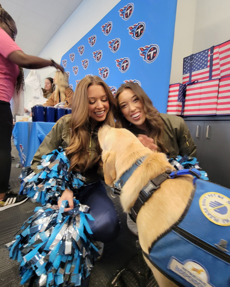 A yellow labrador in a blue service dog vest sniffs two Titans' cheerleaders.They are smiling, looking at the dog and petting him. There is an American flag in the background, and blue and silver pom-poms.
