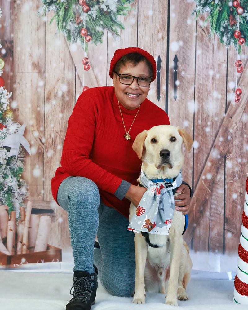 A woman kneels next to a yellow labrador wearing a christmas-themed scarf. They are posed in front of a christmas-themed professional photo backdrop.