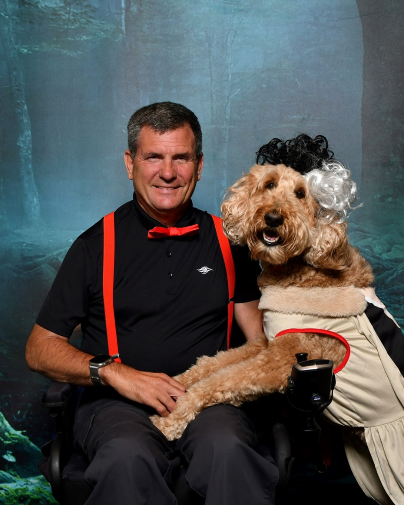 A man in a bowtie sits in a wheelchair in front of a professional photo backdrop. A goldendoodle has its paws in his lap. The dog is wearing a "Cruella de Vil" costume, which has a dress with a fur collar and a black and white wig.