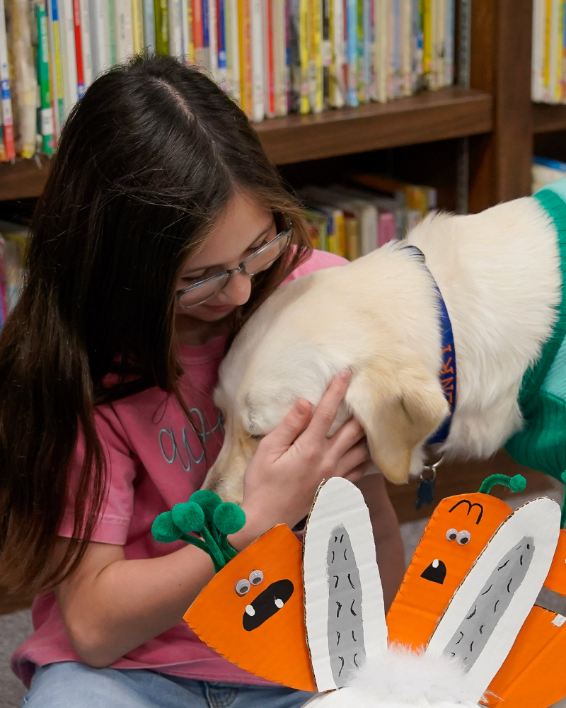 A white labrador in a dog costume leans his head next to a person who is hugging him back.