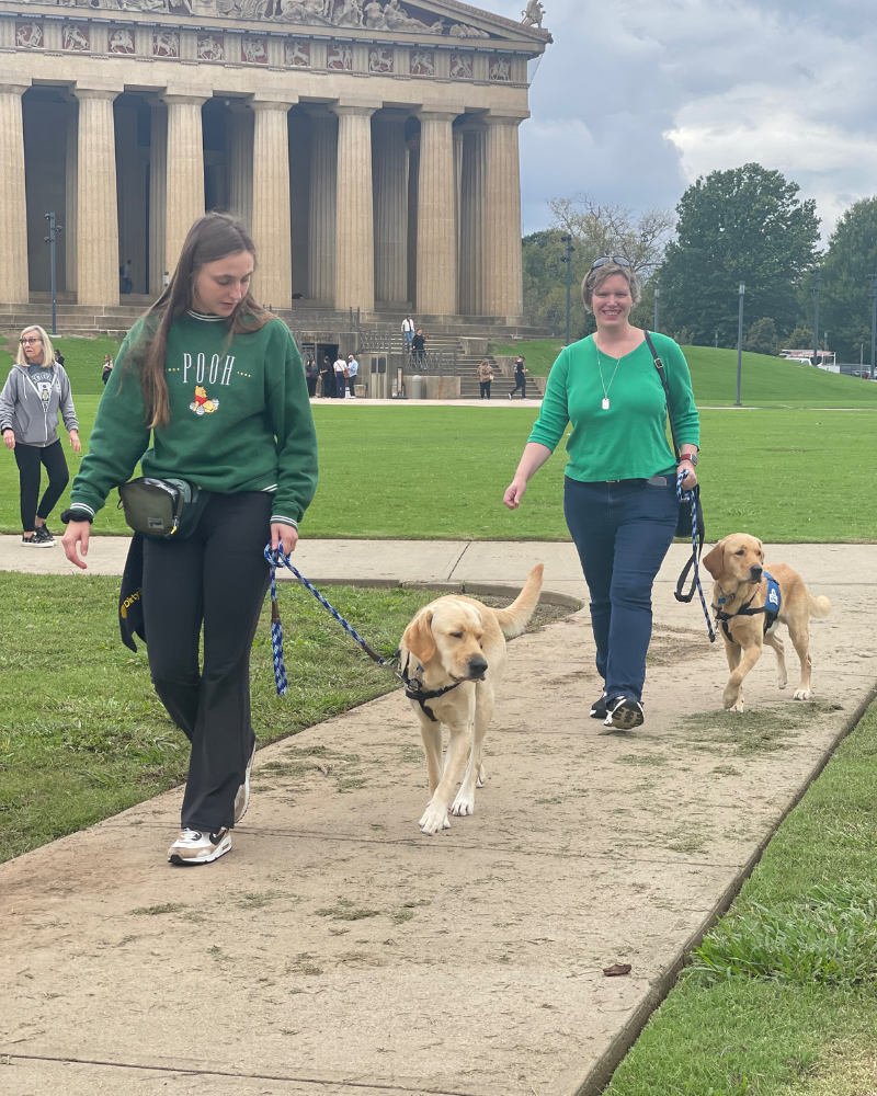 Two women each walk a golden labrador in blue service dog vests on a sidewalk. They are in front of the Parthenon building.