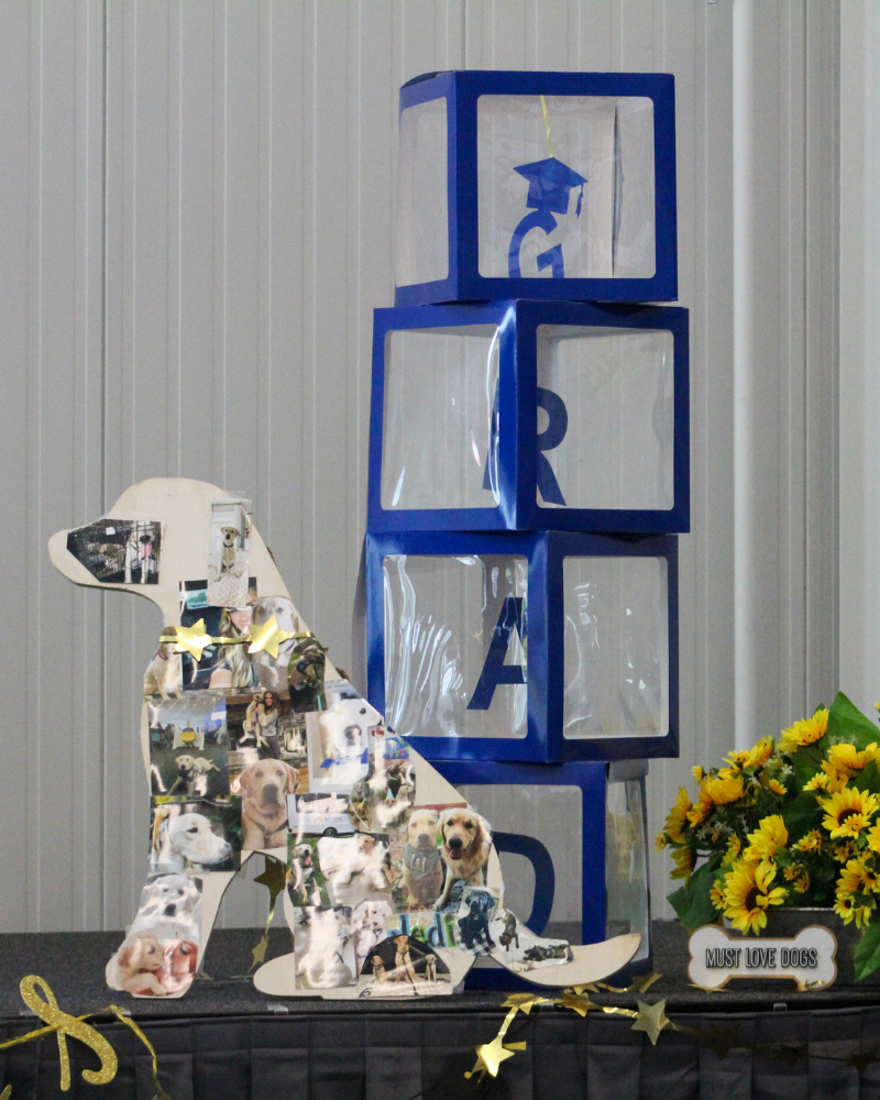 Four plastic blue boxes are stacked on a stage. The contain letters that spell out "GRAD". A wooden stand is next to the boxes. The wooden stand is in the shape of a dog, and has a collage of dog photos within it. There are some golden decorations, stars, a banner, and sunflowers. There is also a decorative sign in the shape of a bone that says "Must Love Dogs".