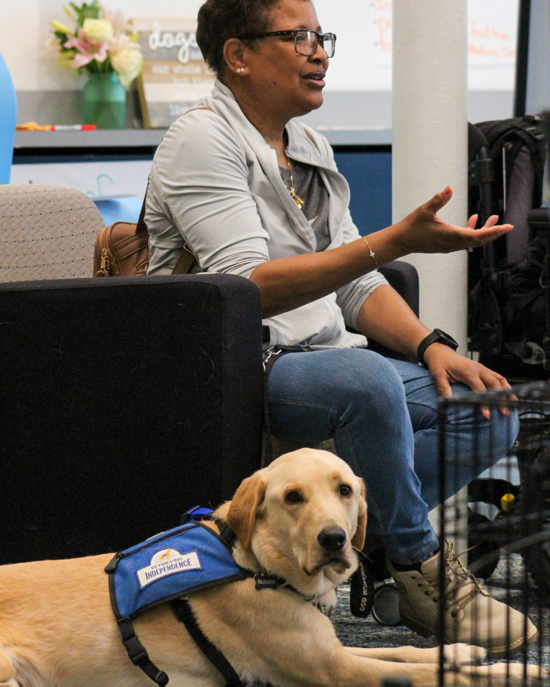 A woman speaks to someone off-camera. At her feet, a golden lab in a blue service dog vest is laying down, looking at the camera.