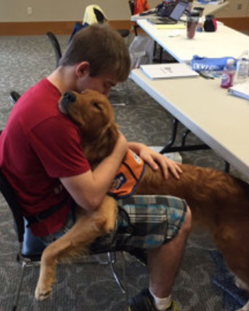 A red golden retriever in an orange service dog vest is in the lap of a young man sitting in a chair. They are hugging each other, with the man's arms around the dog and the dog's head on his shoulder.